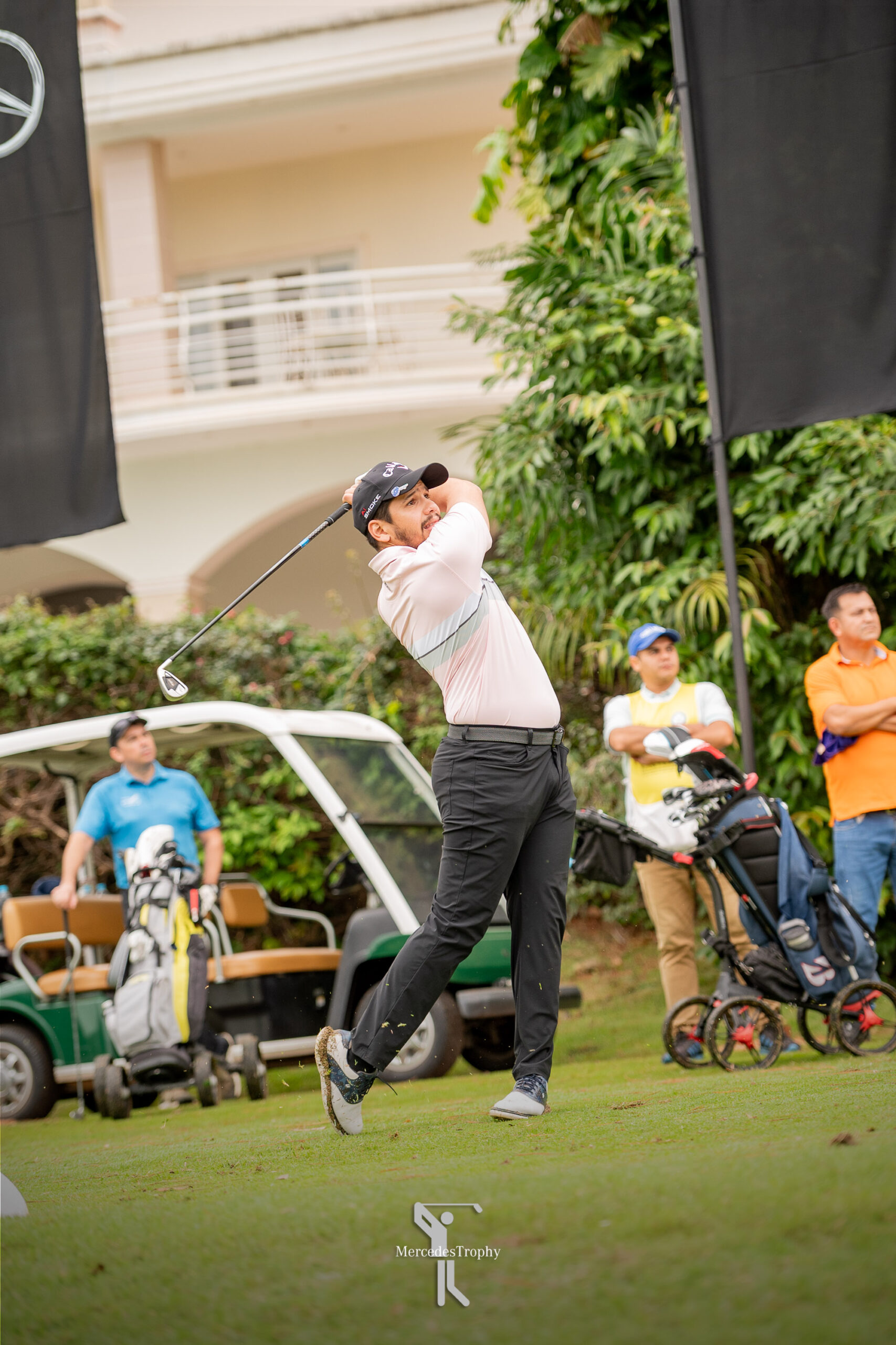 Golfista ejecuta su golpe de salida durante el MercedesTrophy, junto a banderas y carritos de golf.