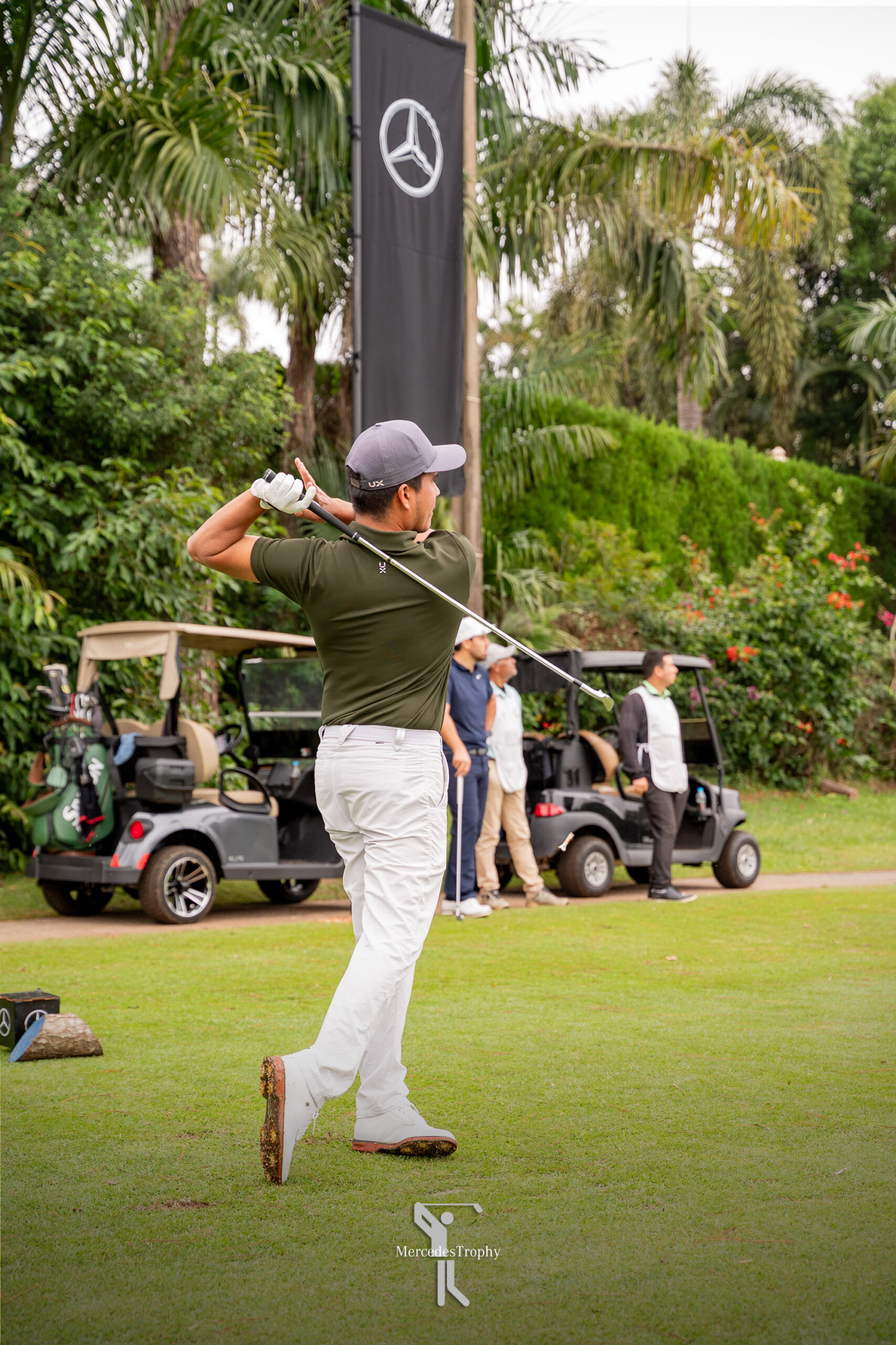 Golfista ejecuta su golpe de salida durante el MercedesTrophy, junto a banderas y carritos de golf.